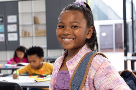 Biracial girl with a purple hair accessory smiles, wearing a backpack in a school classroom. African American boy and another biracial girl focus on schoolwork in the background.の写真素材