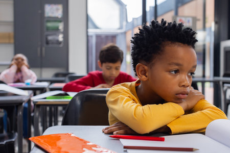 African American boy in a yellow shirt looks sad in a classroom setting with copy space. Biracial boy and girl are focused on their work in the background, highlighting a typical school day.の写真素材