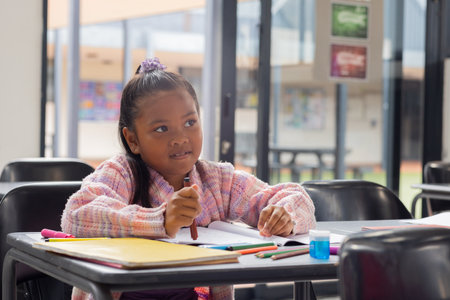 Biracial girl with a focused expression is writing in a notebook at school. Her brown eyes match her hair, tied with a purple accessory, as she studies in a classroom setting.の写真素材