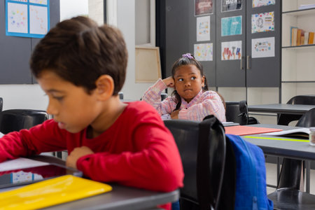 Biracial boy is focused on his work in a school classroom, while a biracial girl looks pensive. They are in a classroom setting, surrounded by educational posters and artwork.の写真素材