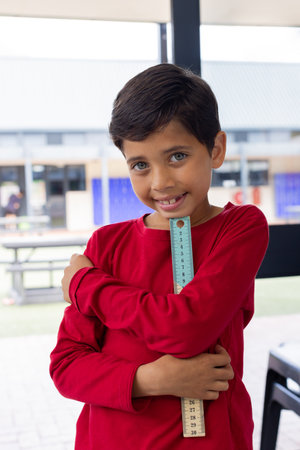 Biracial boy with a bright smile holds a ruler, wearing a red shirt in school. His playful pose...