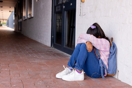 A biracial girl appears distressed, sitting alone with her head on her knees, with copy space. She is outdoors, experiencing a moment of solitude or sadness at school.の写真素材