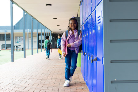 Biracial girl with a bright smile stands by blue school lockers, with copy space. School setting with students in the background, capturing a moment of youthful joy during a break.の写真素材