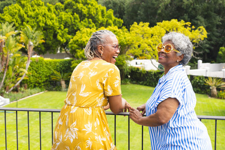 Senior African American woman and senior biracial woman share a joyful moment outdoors. Both are dressed in bright, summery attire, reflecting a warm, casual setting.の写真素材