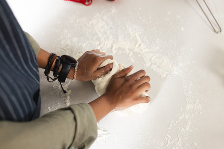 African American young woman kneading dough on table. She wears a striped shirt and bracelets,...