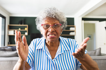 Senior biracial woman with gray hair and glasses looks surprised on a video call. She is indoors,...