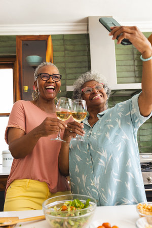 Senior African American woman and senior biracial woman take a selfie with wine glasses. They are in a kitchen, celebrating with joyful expressions and casual attire.の写真素材