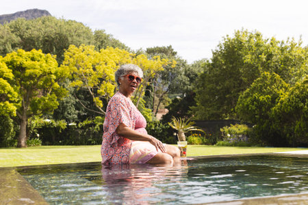 Senior biracial woman enjoys a poolside moment, her gray hair complementing the sunny day. She wears a pink floral outfit, exuding relaxation with a drink in hand, surrounded by lush greenery.の写真素材