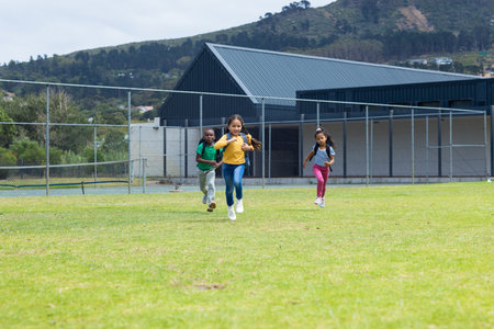 Three children are running joyfully across a grassy field with copy space in school. An African American boy and two girls, one undefined and one biracial, enjoy a playful race outdoors.の写真素材