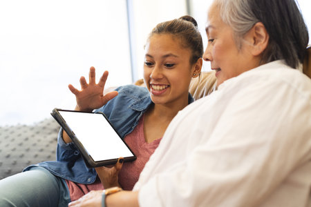 Asian grandmother and biracial teenage granddaughter sitting at home, looking at tablet with copy space. Grandmother has grey hair, granddaughter has light brown hair, unalteredの写真素材