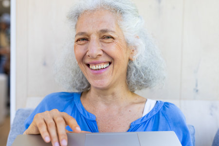 Caucasian senior woman in blue shirt smiles at laptop during home video call. Holding device, sitting at table outside with blue background, enjoying sunny day, unalteredの写真素材