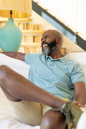 At home, African American senior man laughing on couch. Wearing light blue polo and beige shorts, enjoying a relaxed moment with a cheerful expression, unalteredの写真素材