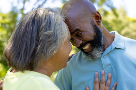 A diverse senior couple laughing outdoors, sporting grey hair. Enjoying sunny day together in park, they're surrounded by a green background, unalteredの写真素材