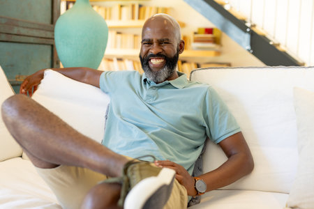 At home, African American senior man wearing light blue polo, smiling broadly. Sitting on a sofa, sporting a bald hairstyle, enjoying a peaceful moment in a cozy atmosphere, unalteの写真素材
