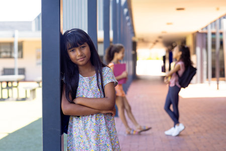 In school, three young biracial female students are standing in the hallway outdoors. They have dark hair, one leaning on a post, others chatting behind, unaltered.の写真素材