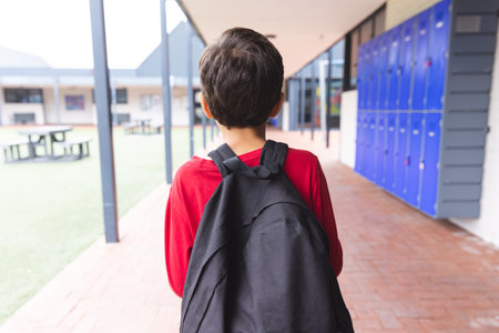 In school, biracial young boy wearing a red shirt and backpack walking outdoors. He has dark hair, and schoolyard has lockers and benches, unaltered.の写真素材