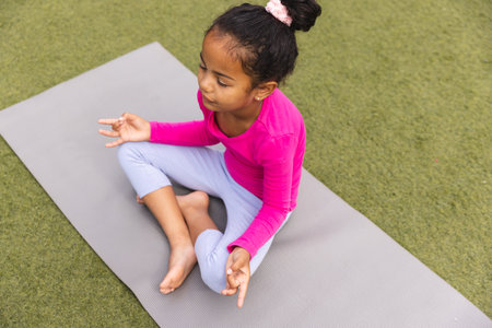 A young biracial girl wearing pink is sitting on a yoga mat outdoors. She has brown eyes, her hair tied up, and is practicing yoga, unaltered.の写真素材