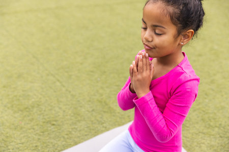 In school, outdoors, a biracial young girl wearing a pink top is practicing yoga on a mat with copy space. She has dark hair tied back, brown eyes, and is concentrating on her pose, unaltered.の写真素材