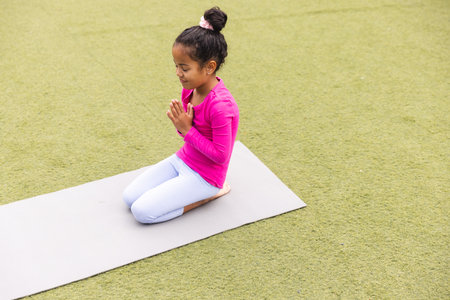 In school, young biracial girl wearing pink is practicing yoga outdoors. She has her hands together, eyes closed, and is sitting on a mat, unaltered.の写真素材