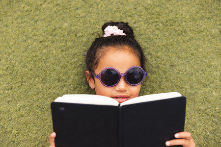 In school, young biracial girl wearing sunglasses is reading a book outdoors. She has dark hair, a pink hair accessory, and a focused expression, unaltered.の写真素材