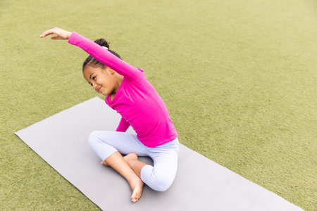 In school, young biracial girl wearing pink and white is stretching on a yoga mat outdoors. She has dark hair tied up, wearing light pants, and practicing on green turf, unaltered.の写真素材
