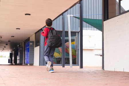 In school, biracial young boy walking along corridor with copy space outdoors. Wearing red hoodie, jeans, and carrying black backpack, he has dark hair, unalteredの写真素材