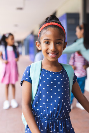 Young biracial girl wearing polka-dot dress and backpack is smiling outdoors. Behind her, a diverse group of students are walking and talking, learning, education, unalteredの写真素材