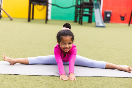 In school, young biracial girl wearing pink is stretching on a mat outdoors doing yoga. She has curly dark hair, a bright smile, and is looking at the camera, unaltered.の写真素材