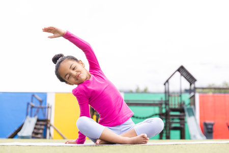 In school, young biracial girl stretching on a mat outdoors doing yoga. She has light brown skin, dark curly hair tied back, and wearing a pink top, unalteredの写真素材