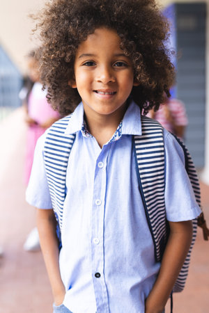 In school, young African American boy with curly hair is smiling outdoors. He wears blue striped shirt and carries a backpack, learning, education, enjoying his day, unalteredの写真素材