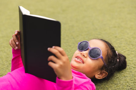 In school, young biracial girl wearing sunglasses lying on grass outdoors reading. She has dark hair in braids, a bright pink top, and is enjoying her book, unaltered.の写真素材