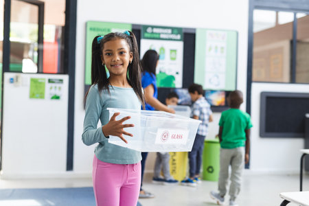 In a school classroom, diverse students and a teacher focus on recycling. The young girl in front holds a bin, ready to sort glass, learning, education, unalteredの写真素材