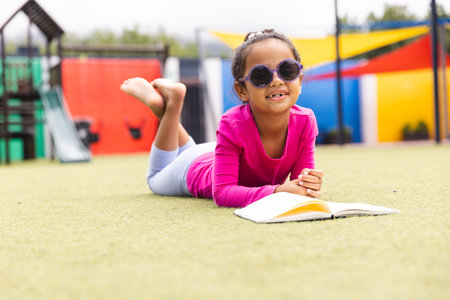In school, outdoors, a biracial young girl wearing sunglasses is reading. She has brown skin, dark curly hair, and is dressed in pink, unaltered.の写真素材
