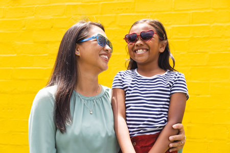 In front of a bright yellow wall outdoors, a biracial young girl and a woman are smiling. Both wearing sunglasses, the girl has heart-shaped frames and the woman has blue striped ones, unaltered.の写真素材
