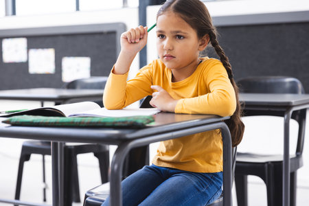 In school, young biracial girl sitting at a desk in a classroom, looking thoughtful. Wearing a yellow top, she has long brown hair and dark eyes, unalteredの写真素材