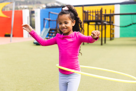 In school, outdoors, a biracial young girl is playing with a ring hoop. She has light brown skin, dark curly hair, and is wearing a pink top.の写真素材