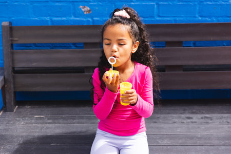 In school, young biracial girl wearing pink blowing bubbles outdoors. She has curly dark hair, light brown skin, and is sitting on a bench, unaltered.の写真素材