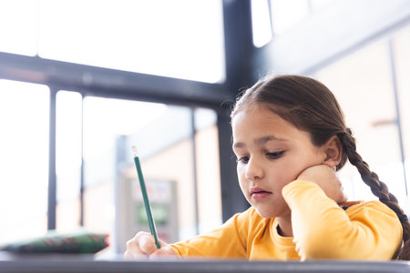 In school, in the classroom, a young biracial girl with braided hair is writing with copy space. She wears a yellow top and focuses on her work, surrounded by math signs, unaltered.の写真素材