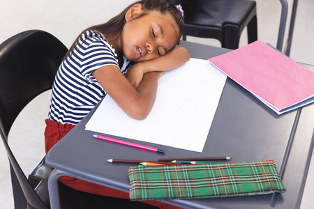 In school, young biracial girl wearing a striped shirt resting head on desk in classroom. She has brown hair, light brown skin, and is surrounded by school supplies, unaltered.の写真素材