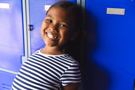 In school, young biracial girl with a bright smile standing by blue lockers. She has long dark hair, wearing a striped shirt, and looking happy, unalteredの写真素材