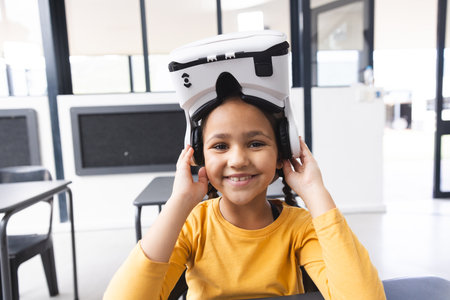 In school, in the classroom, a young biracial girl wearing a VR headset is smiling. She has dark hair, brown eyes, and is in a yellow shirt, unaltered.の写真素材