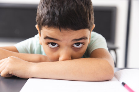 In school, young biracial boy leaning on desk in classroom, looking bored. He has dark hair, brown eyes, wearing a light green t-shirt, unaltered.の写真素材