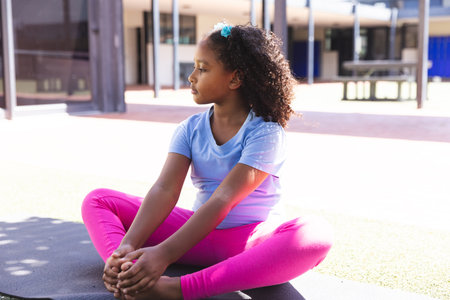 Biracial girl sits cross-legged outdoors at school. She appears relaxed in a school setting, enjoying a sunny day.の写真素材