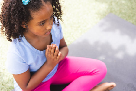Biracial girl practices yoga outdoors at school, with copy space. She sits on a mat in a peaceful pose, promoting health and mindfulness.の写真素材