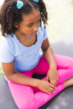 Biracial girl sits on a yoga mat outdoors at school. She appears focused and calm in a tranquil...
