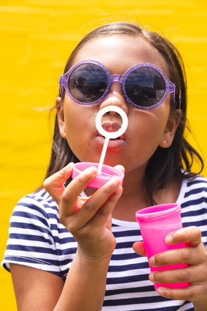 In school, young biracial girl wearing sunglasses is blowing bubbles outdoors. She has dark hair, wearing a striped shirt against a yellow background, unaltered.の写真素材