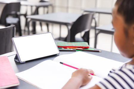 In school, a young biracial girl is writing in a notebook in the classroom, with copy space. She has a tablet with a blank screen on the desk, unaltered, with copy space.の写真素材