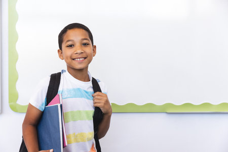 Biracial boy ready for school, with copy space. He stands smiling with his backpack and books, signaling a new day of learning.の写真素材