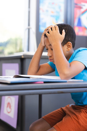 Biracial boy looks frustrated with schoolwork in the classroom at school. He is concentrating on his...