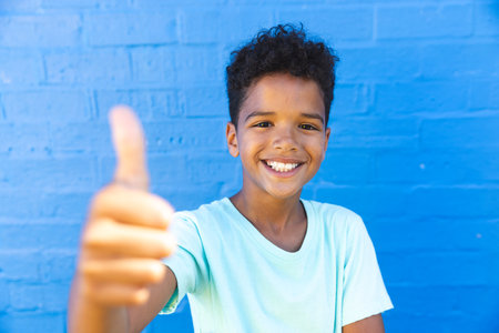 Biracial boy gives a thumbs up against a blue wall. His bright smile and gesture exude confidence and positivity.の写真素材